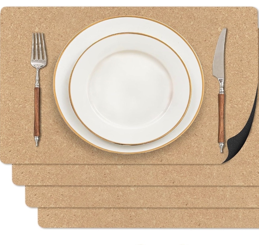 A set of four easy corkboard placemats with a fork, knife, and white plates with gold rims arranged on the top mat, showing a simple table setting. The top right corner of the placemat is slightly lifted.